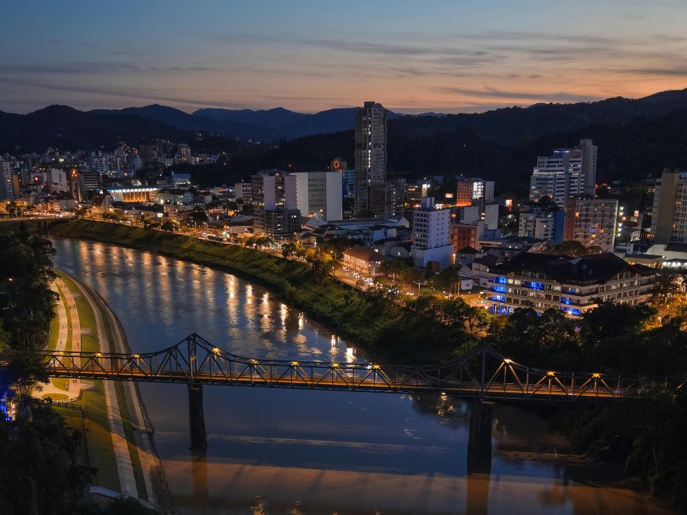 Paisagem noturna de Blumenau com rio iluminado e ponte metálica, ótima opção para conteúdos sobre O que fazer em Blumenau à noite.